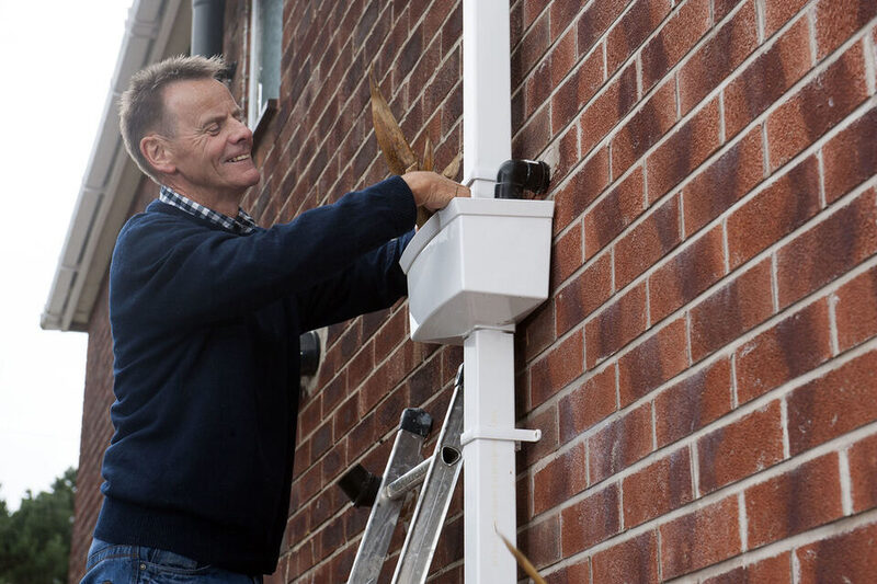 Man on a ladder clearing leaves from a white gutter downpipe
