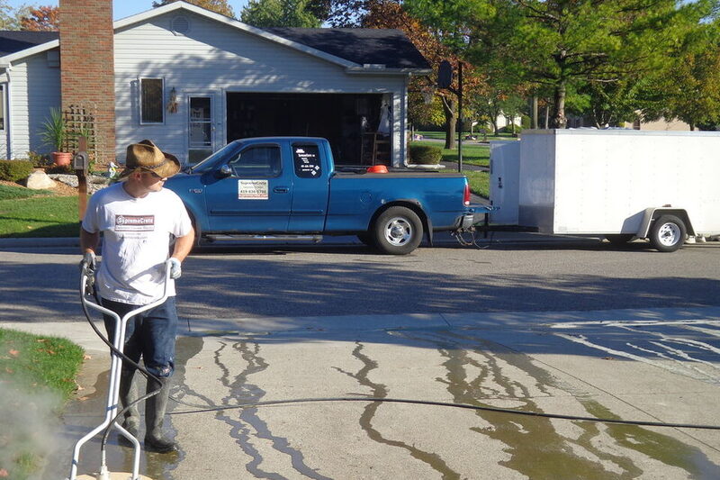 Worker water blasting a residential driveway with work ute and trailer